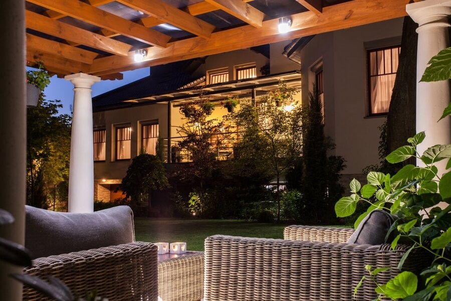 A covered patio setup in a backyard during the evening featuring seats and lighting fixtures.