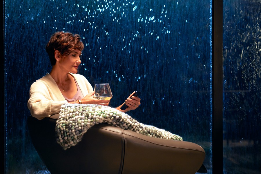 Woman sitting with a glass of wine and her phone with a rainy night in the windows behind her. 