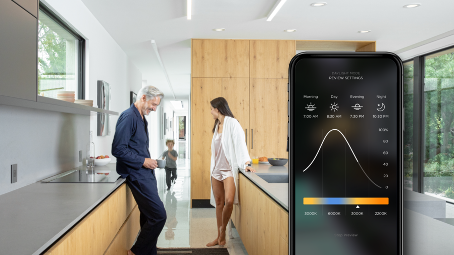 A couple having a conversation in the kitchen while their young child runs toward them. A phone with the Savant app displays the lighting settings.