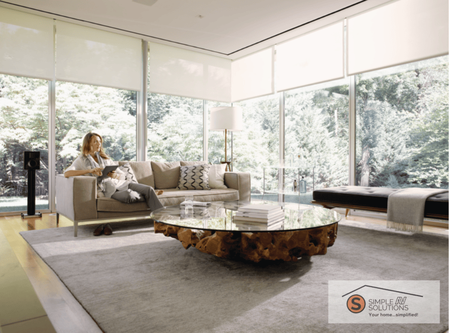 A woman sitting in a living room with floor-to-ceiling windows that have half-drawn motorized shades.