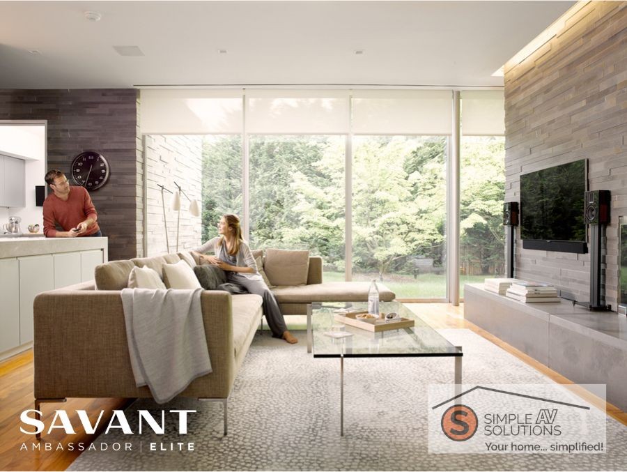 A woman sits on the couch in the living room while talking to a man in the kitchen. Window shades are partially lowered.