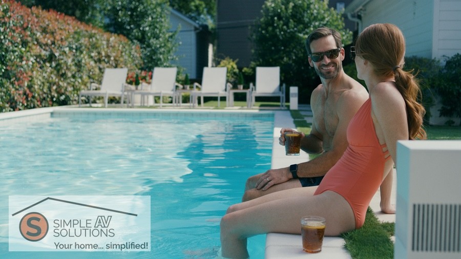 A man and woman relax by a pool, each holding a drink. Chairs are visible in the background, surrounded by landscaping.