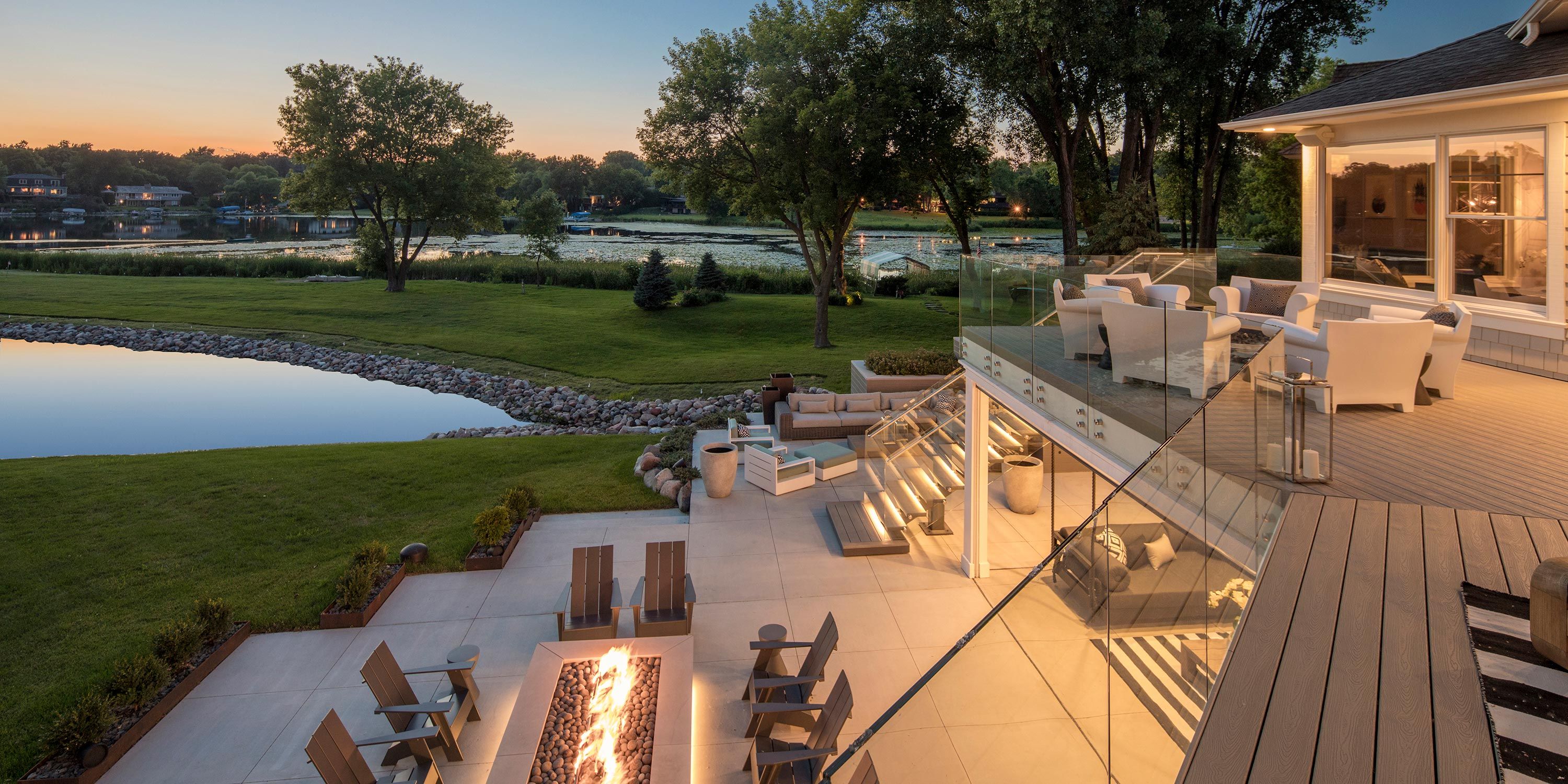 A serene backyard with a pond, lush green lawn, and outdoor seating areas overlooking the water at sunset.