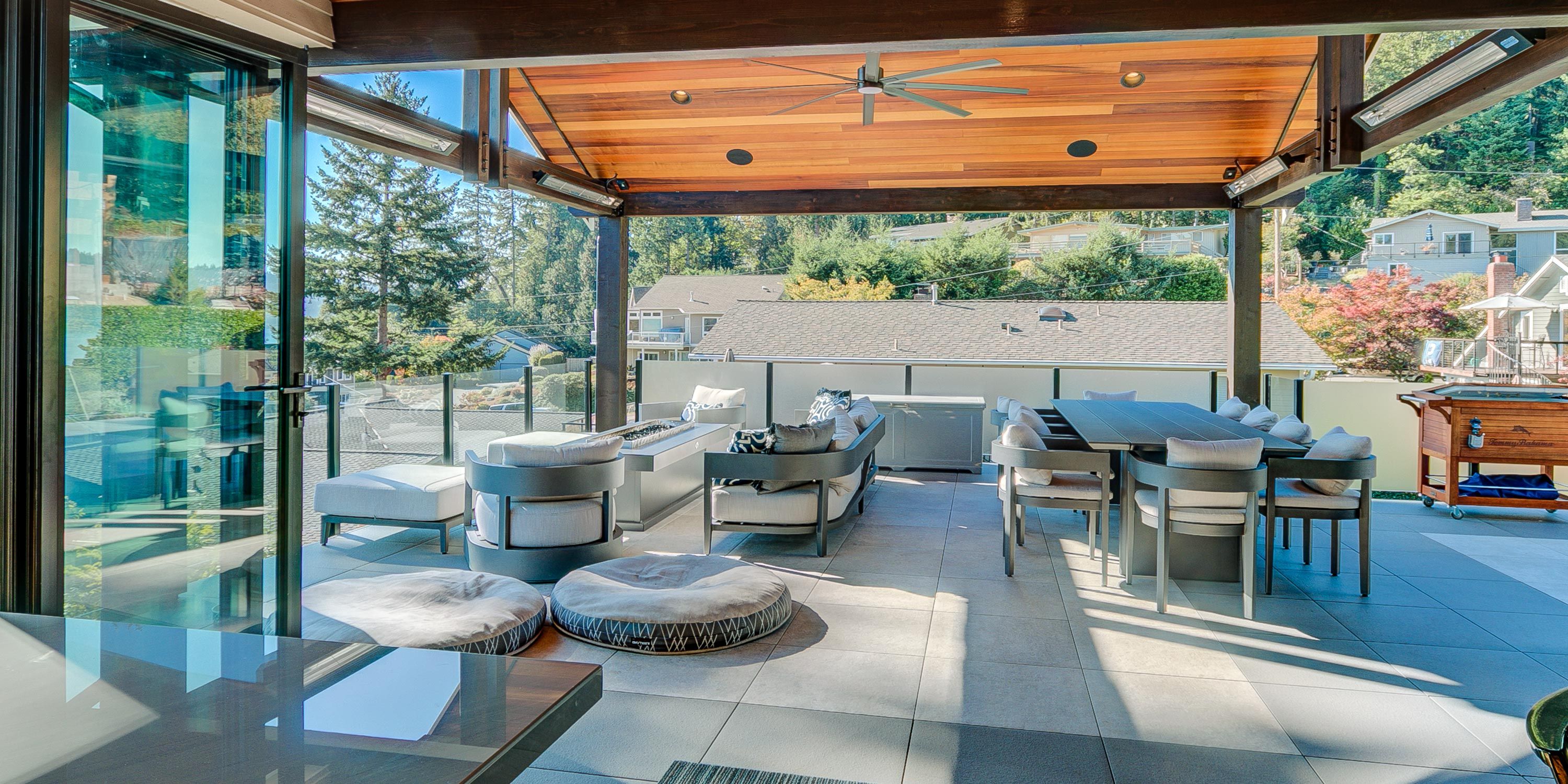 A spacious covered outdoor patio with modern seating, a large dining table, and views of a suburban neighborhood, featuring a wood-paneled ceiling, ceiling fan, and integrated heating elements.