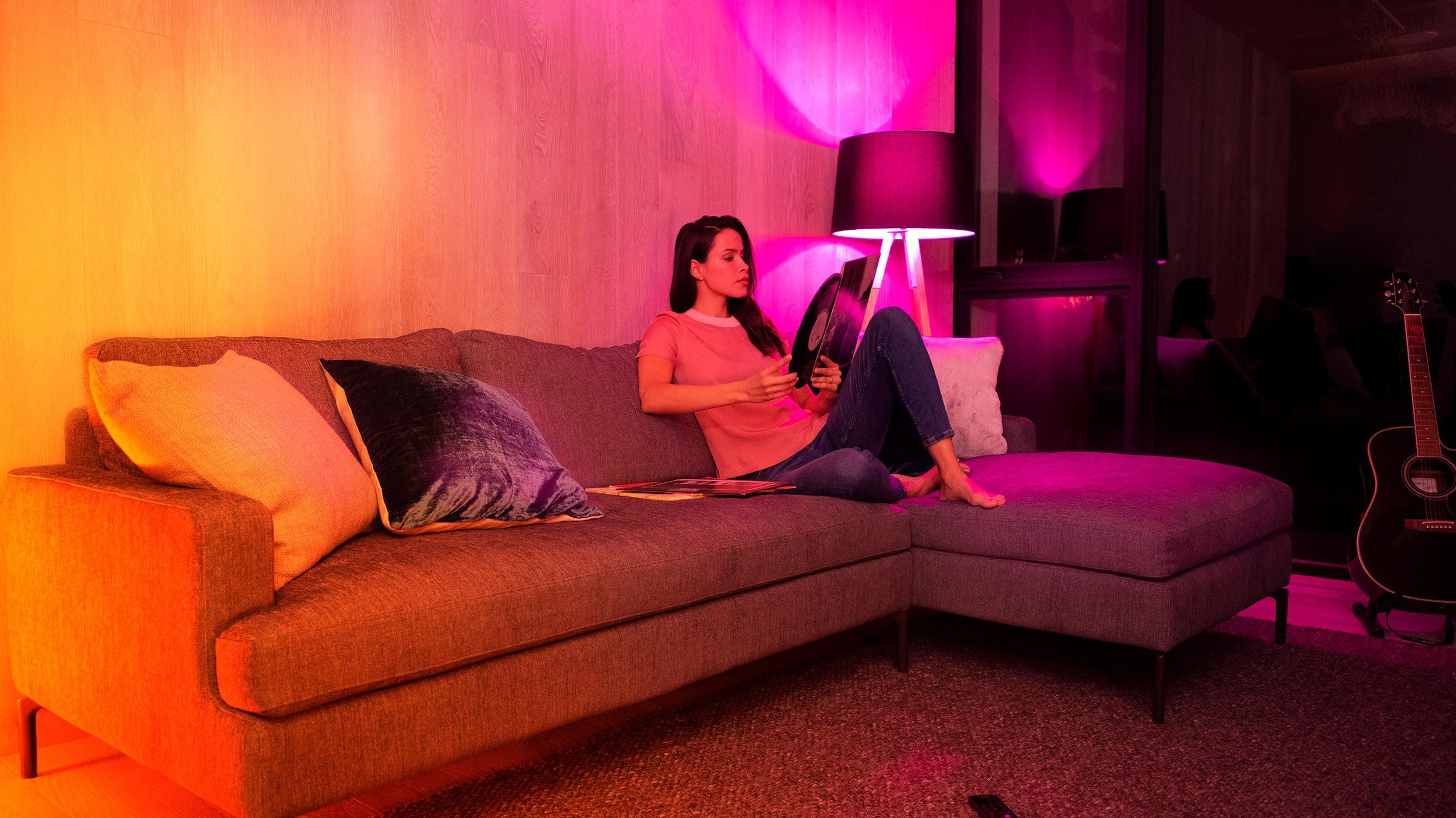 A woman relaxing on a modern sectional sofa, illuminated by vibrant colored lighting, while holding a vinyl record, creating a cozy and artistic atmosphere.