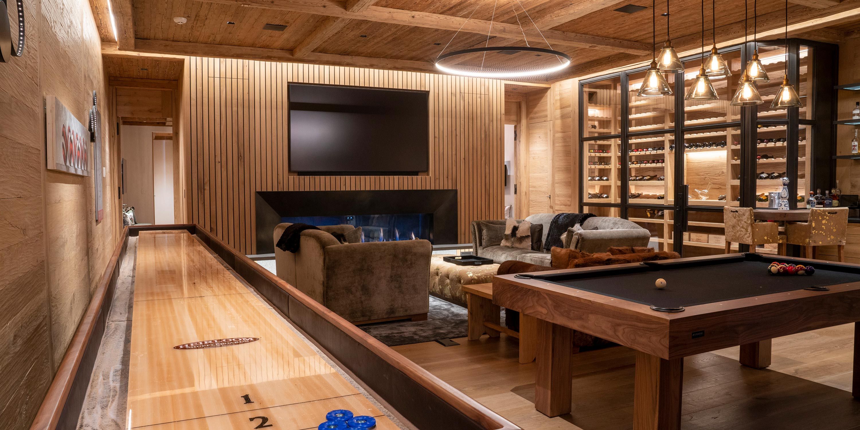 A cozy game room featuring a shuffleboard table, pool table, plush seating, and a wine display, creating a luxurious entertainment space.