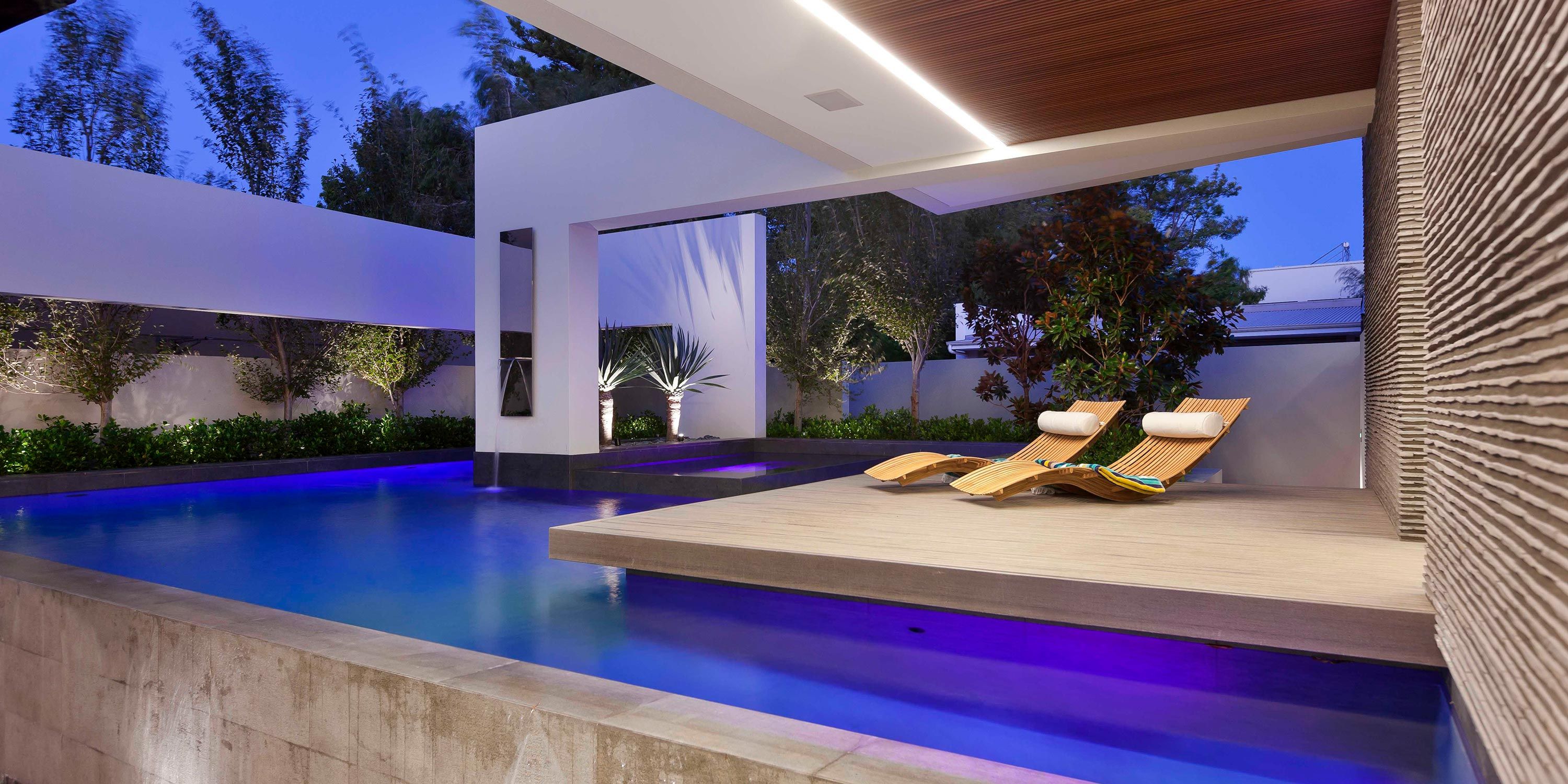 A luxurious pool area with modern lounge chairs, a tranquil water feature, and ambient lighting, set against a backdrop of evening sky.